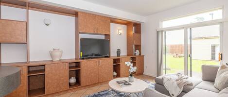 A modern living room with a built-in TV, wooden cabinets, a grey sofa, and a large sliding glass door to the outdoors.