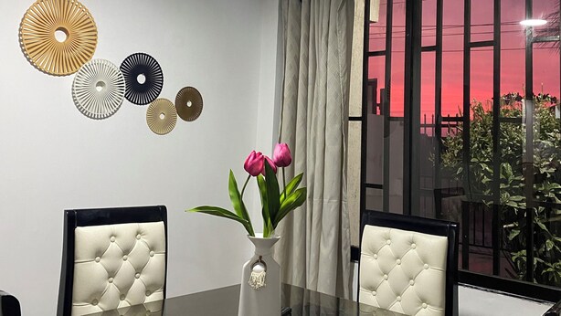 Un comedor moderno con una mesa de vidrio, dos sillas tapizadas, un jarrón con flores y decoración en la pared.
