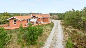 A single-story brick house with a tiled roof, a patio with outdoor furniture, and a gravel driveway leading to a forested area.