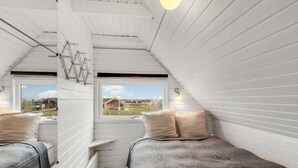 Una habitación pequeña con paneles blancos, dos camas, una ventana con vistas a un paisaje rural y un techo inclinado.