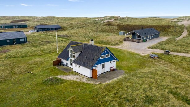 Una casa costera con techo inclinado, rodeada de campos verdes y dunas, que cuenta con un pequeño patio y un asta de bandera visible.