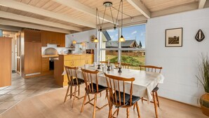 Una cocina moderna con armarios de madera y una isla grande, un comedor con mesa blanca y sillas de madera, y vista a una casa a través de amplios ventanales.