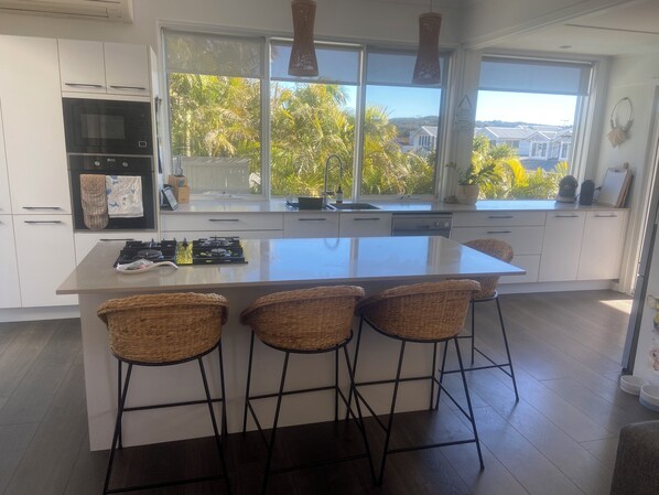 Una cocina moderna con una isla grande, muebles blancos y vistas a palmeras a través de amplias ventanas.