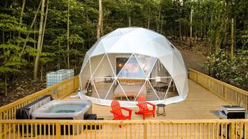 Une structure en forme de dôme géodésique, dotée d’une terrasse en bois, de deux chaises rouges, d’un jacuzzi et d’une petite table.