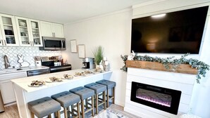 A modern kitchen with a central island, bar stools, a flat-screen TV, a fireplace, and a patterned rug.