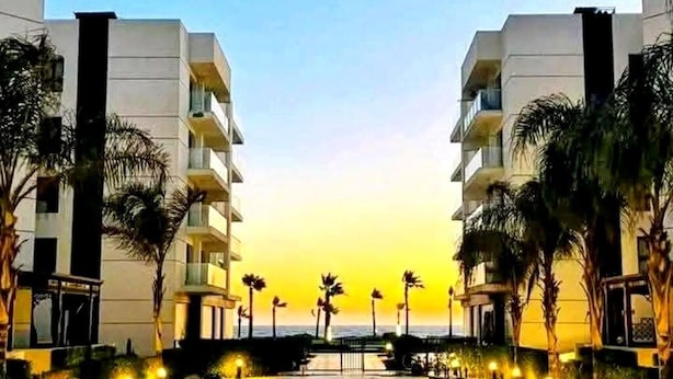 Un hotel con un camino central bordeado de palmeras e iluminado con luces en el suelo, que conduce a un edificio con balcones.