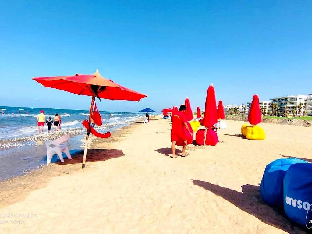 A beach with red umbrellas, a white plastic chair, and a blue bag on the sand.