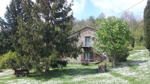 Ein Steinhaus mit einem Balkon, umgeben von Bäumen und einer Rasenfläche mit weißen Blumen.