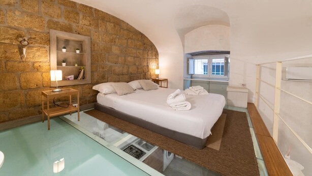 A bedroom with a stone wall, a bed with white linens, a bedside table with a lamp, and a window with a view of the outside. - Vico Santo Spirito di Palazzo 34 (Naples)