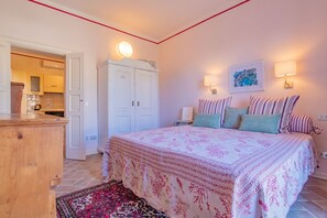 A bedroom with a bed, a nightstand, a dresser, and a kitchen visible through an open door.