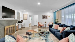 Una sala de estar moderna con un sofá azul oscuro, una mesa de centro de vidrio y una alfombra colorida. Hay un televisor de pantalla plana montado en la pared, una mesita con una lámpara y una puerta que da a otra habitación.
