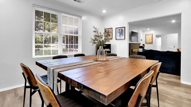 Un comedor con una mesa de madera, sillas y un jarrón con flores. Se aprecia una ventana con persianas, un cuadro en la pared y una zona de cocina al fondo.