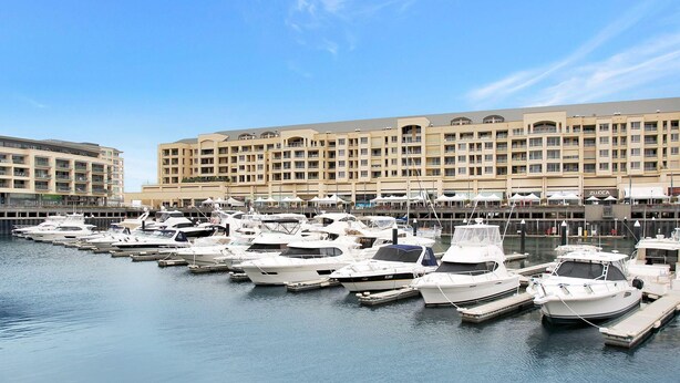 A marina with multiple boats docked in front of a large hotel with multiple balconies and a flat roof. - Belle Escapes - Glenelg Seaside Apartment (Glenelg)