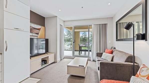 A modern living room with a flat-screen TV, a coffee table, a sofa, and a balcony with a view of trees.