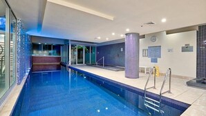 A swimming pool with clear blue water, a tiled deck, and a glass wall on one side.