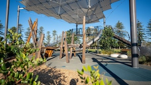 Kids can play on the slide and climbing elements in this playground area surrounded by a fence and shaded by trees.