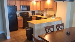 A modern kitchen with wooden cabinets, a black refrigerator, a microwave, and a coffee maker. There is a dining area with a wooden table and chairs. - Beautiful 1 bedroom condo on the beach in Pompano Beach (Pompano Beach)