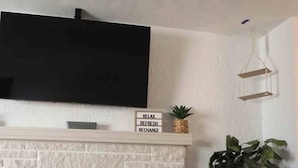 A living room with a white brick fireplace, a black TV mounted above it, and a small shelf with motivational words. - Luxury 5 Bedroom Ranch Home: Walk to Notre Dame Campus / 2 Full Baths +Game Room (South Bend)