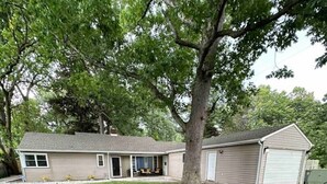 A single-story house with a garage, a covered porch, and a large tree in front. - Luxury 5 Bedroom Ranch Home: Walk to Notre Dame Campus / 2 Full Baths +Game Room (South Bend)