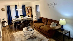 A living room with a brown leather sofa, a round coffee table, a dining table with blue curtains, and a 'Be Our Guest' sign on the wall. - Luxury 5 Bedroom Ranch Home: Walk to Notre Dame Campus / 2 Full Baths +Game Room (South Bend)