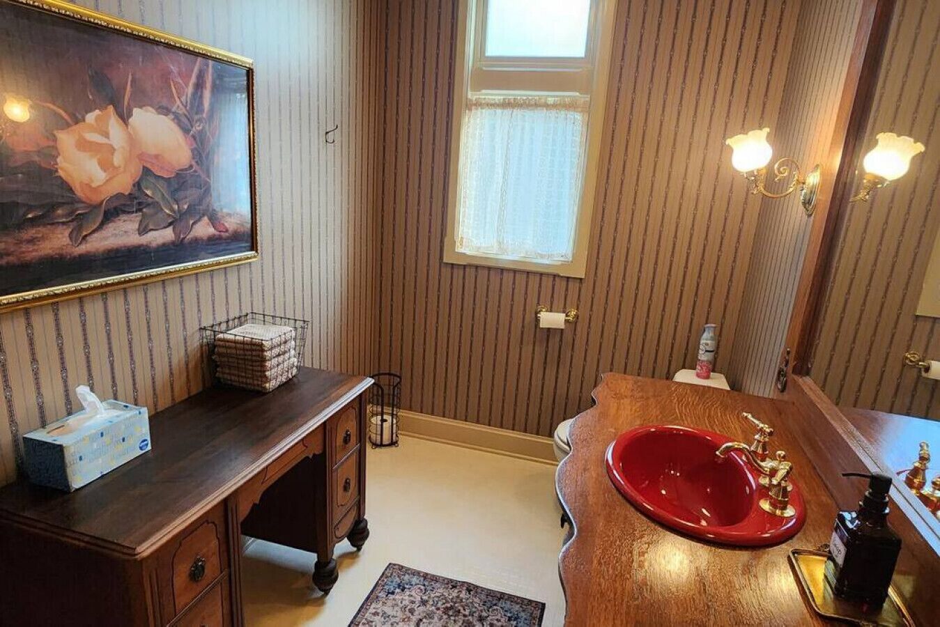A bathroom with a wooden vanity, a red sink, a framed floral painting, and a window with vertical blinds.