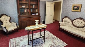 A room with a wooden cabinet, two upholstered chairs, a table with a lace tablecloth, and a patterned rug.