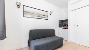 Een kamer met een grijze bank, een lijst met een landschap, een keuken met een blauwe achterwand en een witte deur.