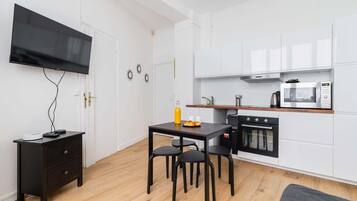 Een compacte keuken met witte kastjes, een magnetron, een oven, een kleine eettafel met stoelen, een televisie aan de muur en een zwart nachtkastje met lades.