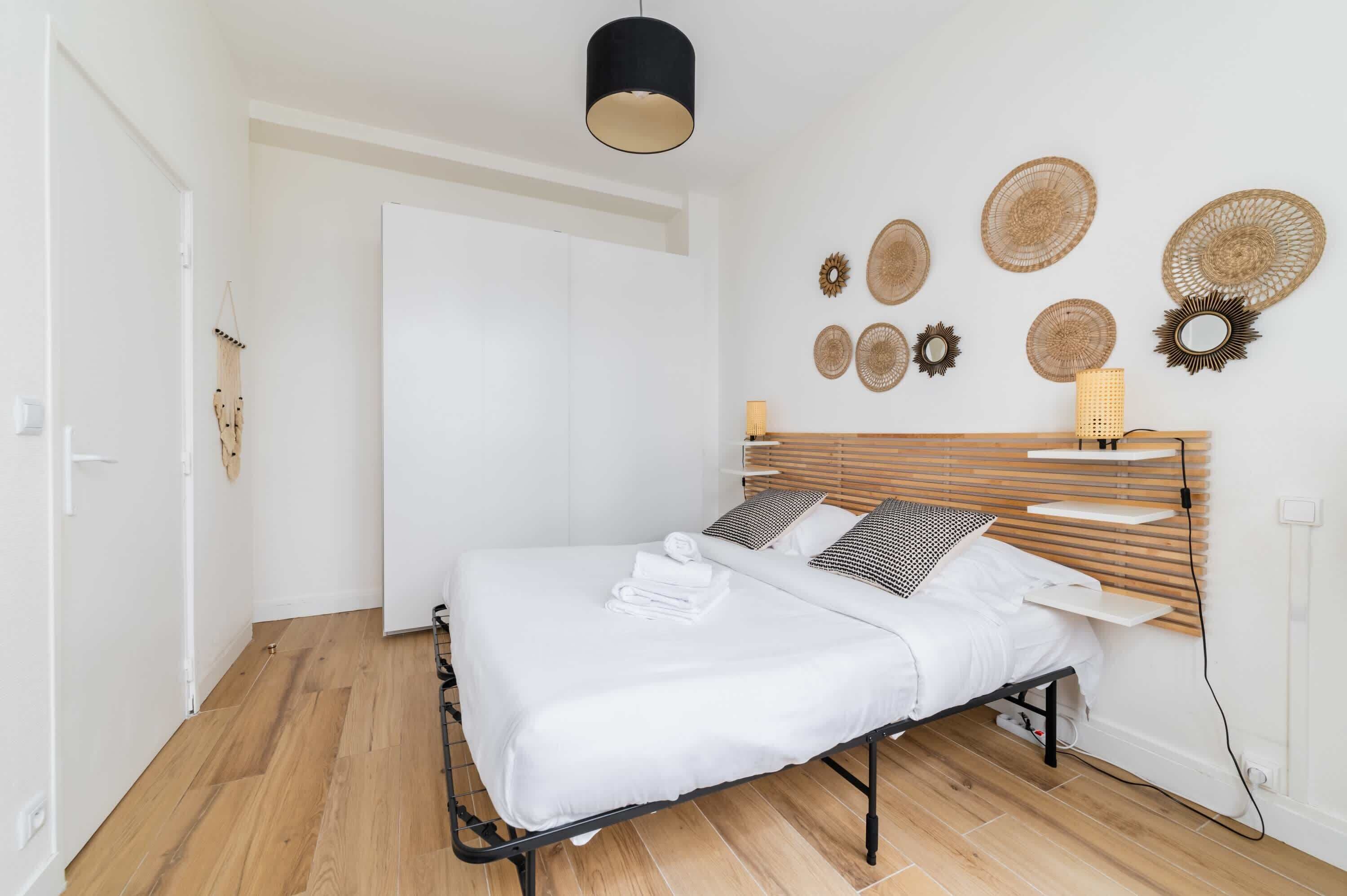A neatly made bed with white linens and a wooden headboard, a wall-mounted lamp, and several woven wall decorations.