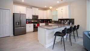 Una cocina moderna con armarios blancos, electrodomésticos de acero inoxidable, una isla central con taburetes y luces colgantes.