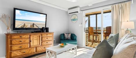 A living room with a television, wooden cabinet, white coffee table, teal sofa, and a view through a glass door to an outdoor deck.