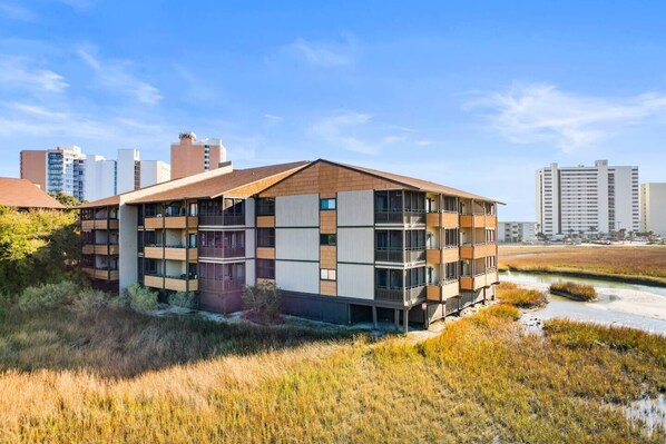 A multi-story building with balconies, situated near a water body with vegetation.