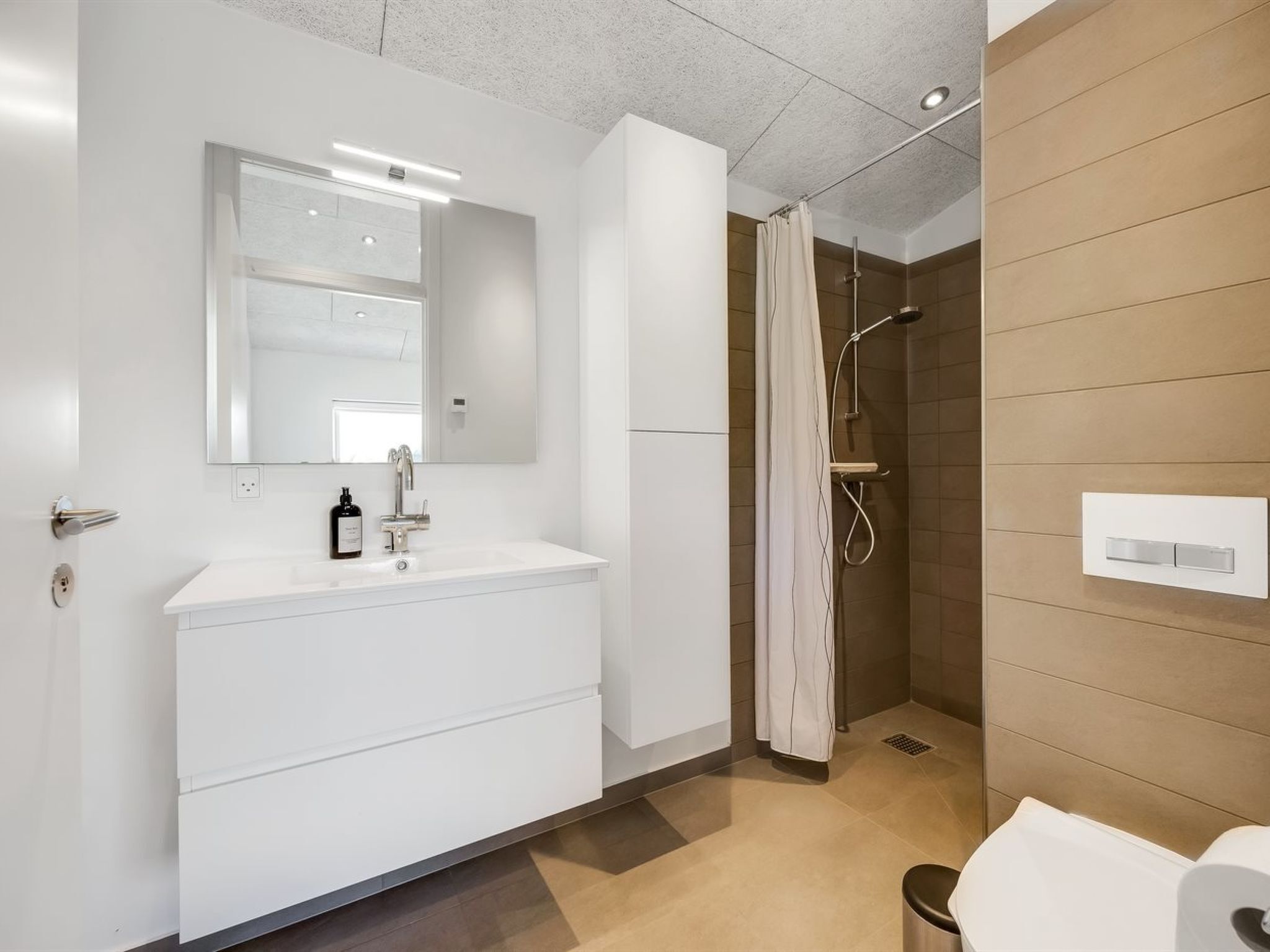 Un baño moderno con un tocador blanco, un espejo grande, una ducha con cortina y un interruptor de luz en la pared.