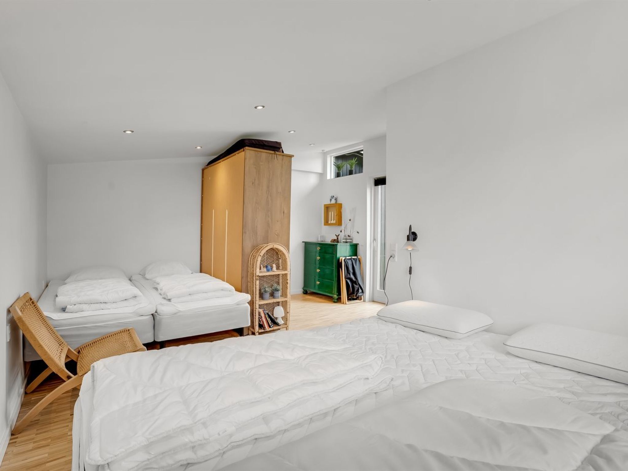 Une chambre moderne avec deux lits, une armoire en bois, une commode verte et un fauteuil en osier.