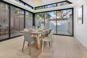 A dining area with a large table set for six, surrounded by chairs, in a modern house with large windows and a view of the backyard.