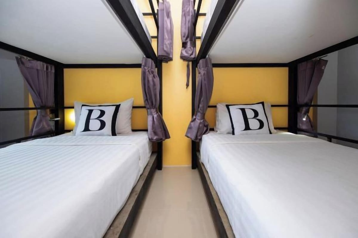 A hotel room with two beds, each with a pillow featuring a large letter, a window with curtains, and a ceiling with hanging fabric.