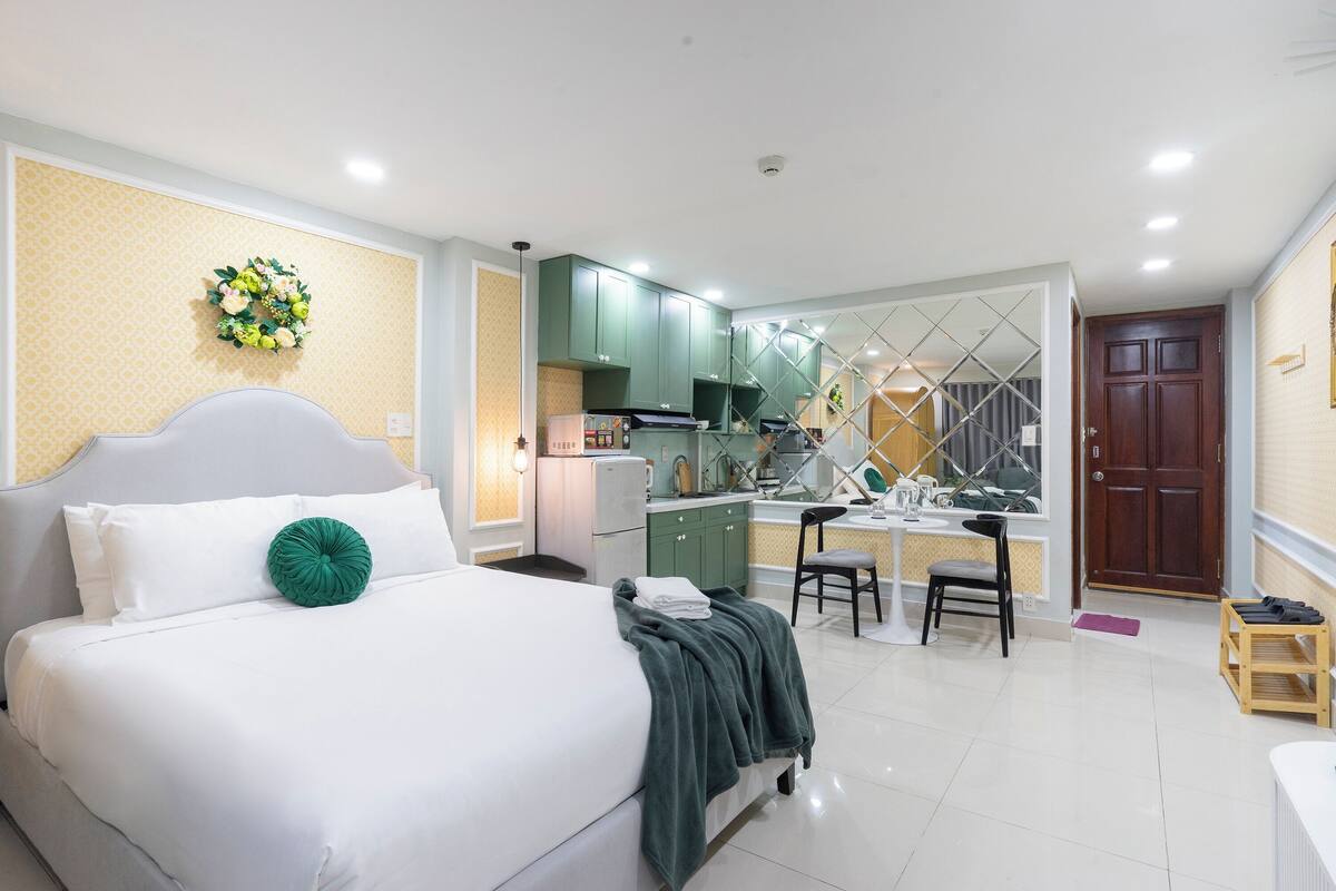 A bedroom with a bed, a green pillow, a kitchen area with a dining table and chairs, and a wooden door.