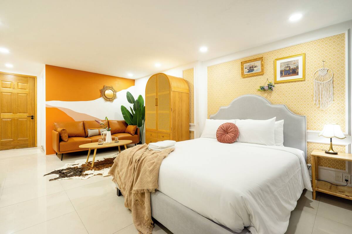 A bedroom with a large bed, a sofa, a wooden wardrobe, and a nightstand with a lamp.