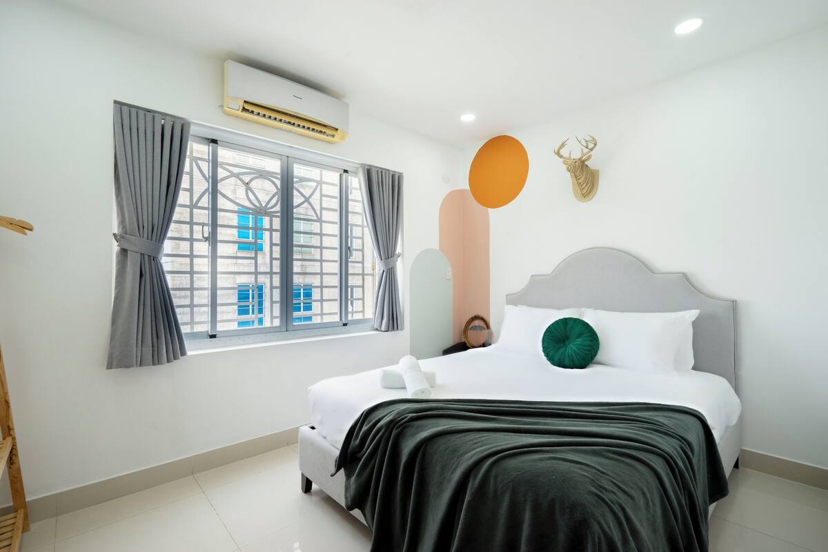 A modern hotel room with a large bed, a window with curtains, a mounted air conditioner, and decorative wall art.
