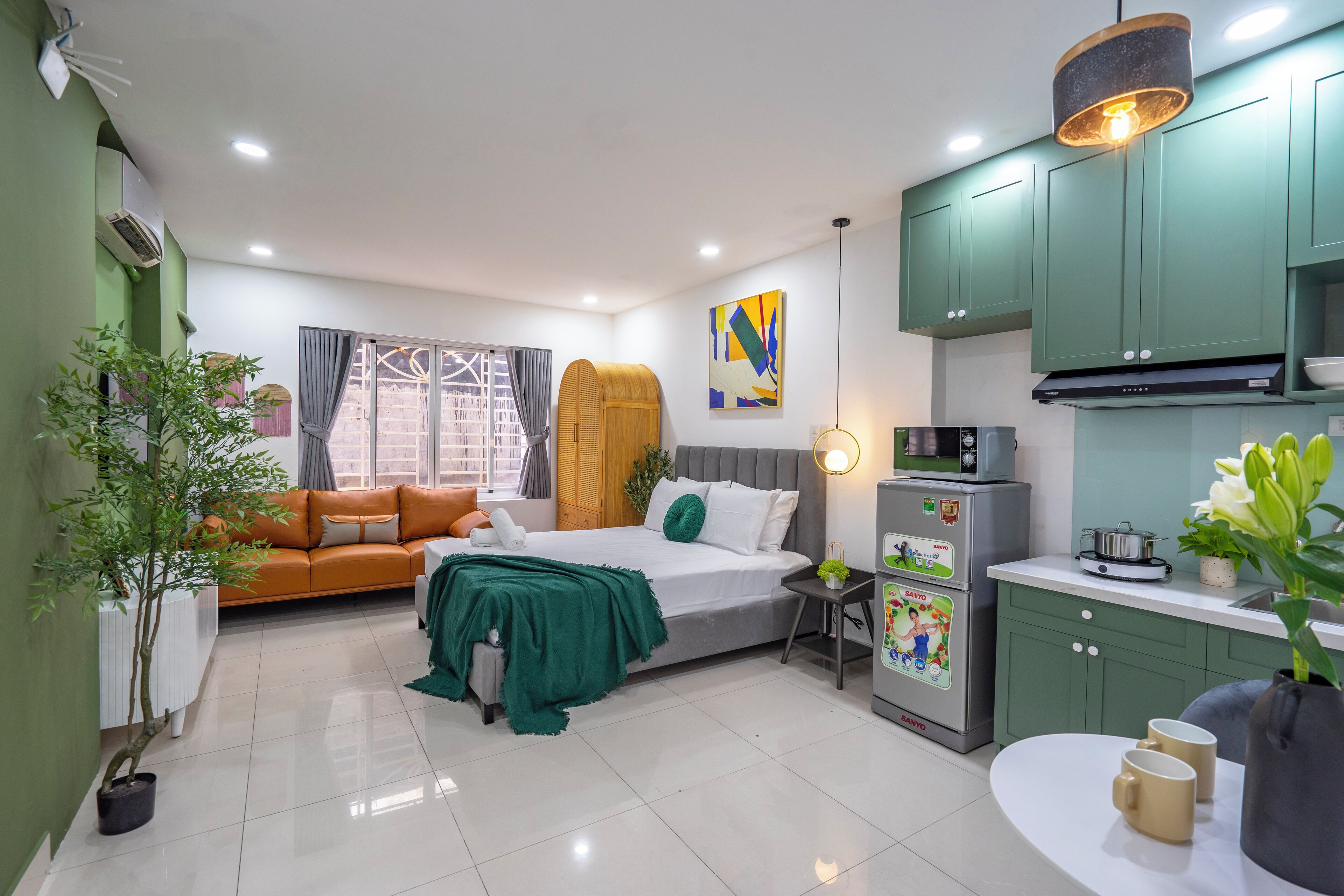 A compact hotel room with a bed, a sofa, a small kitchen area, and a potted plant.