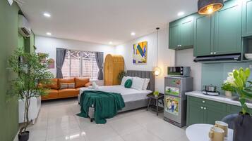 A compact hotel room with a bed, a sofa, a small kitchen area, and a potted plant.