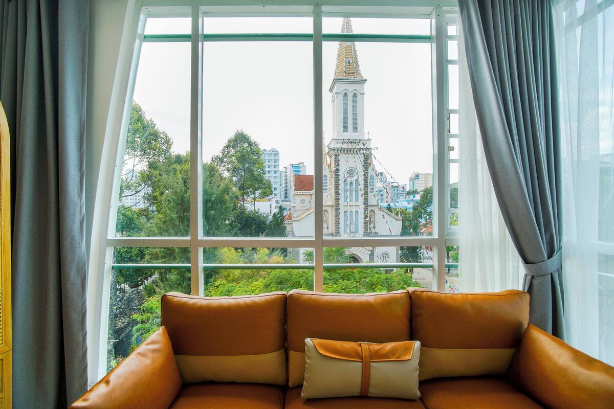 A room with a large window, a brown leather sofa, and a view of a church with a tall tower.