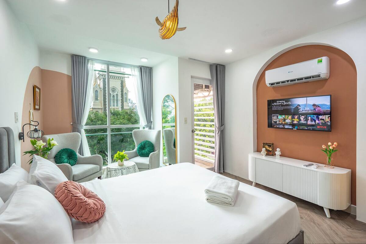 A modern hotel room with a large bed, a TV mounted on the wall, a balcony with a view of a building, and a ceiling fan.