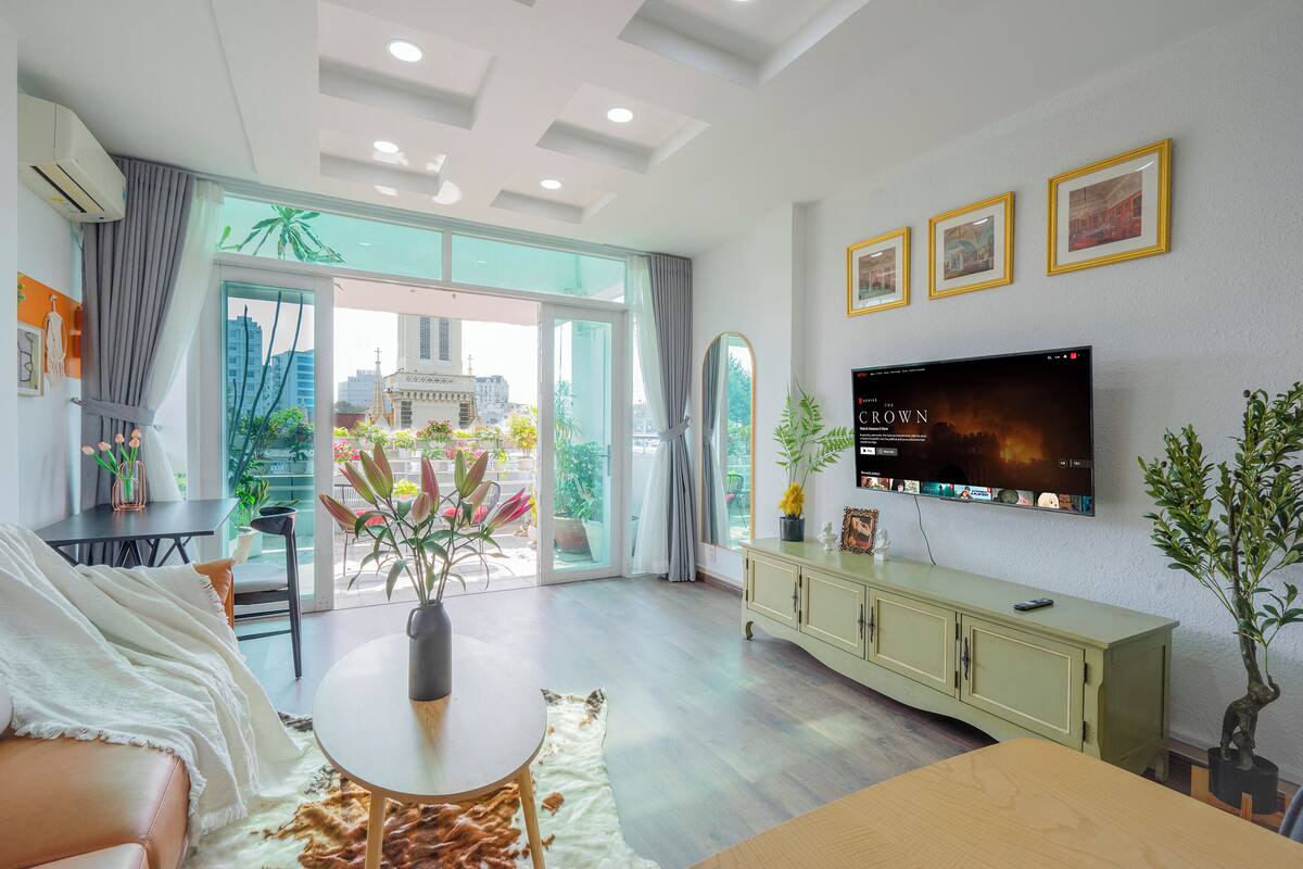 A modern living room with a large TV, a dining area, and a view of the cityscape through the glass doors.