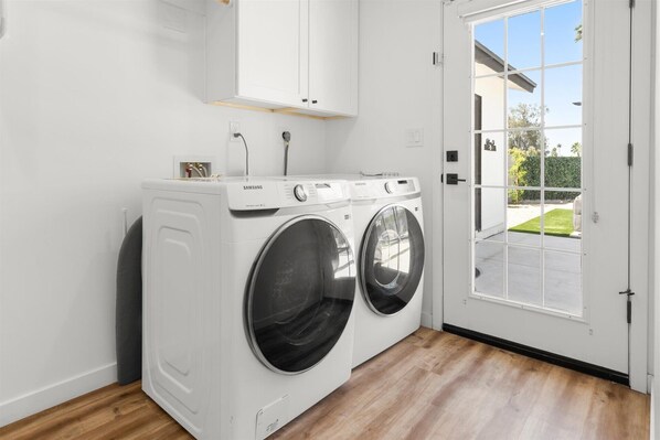 A laundry room with two white Samsung front-loading washers, a wooden floor, and a door leading outside. - New Paradise Listing - Resort Pool, Putting, Game Room (Scottsdale)