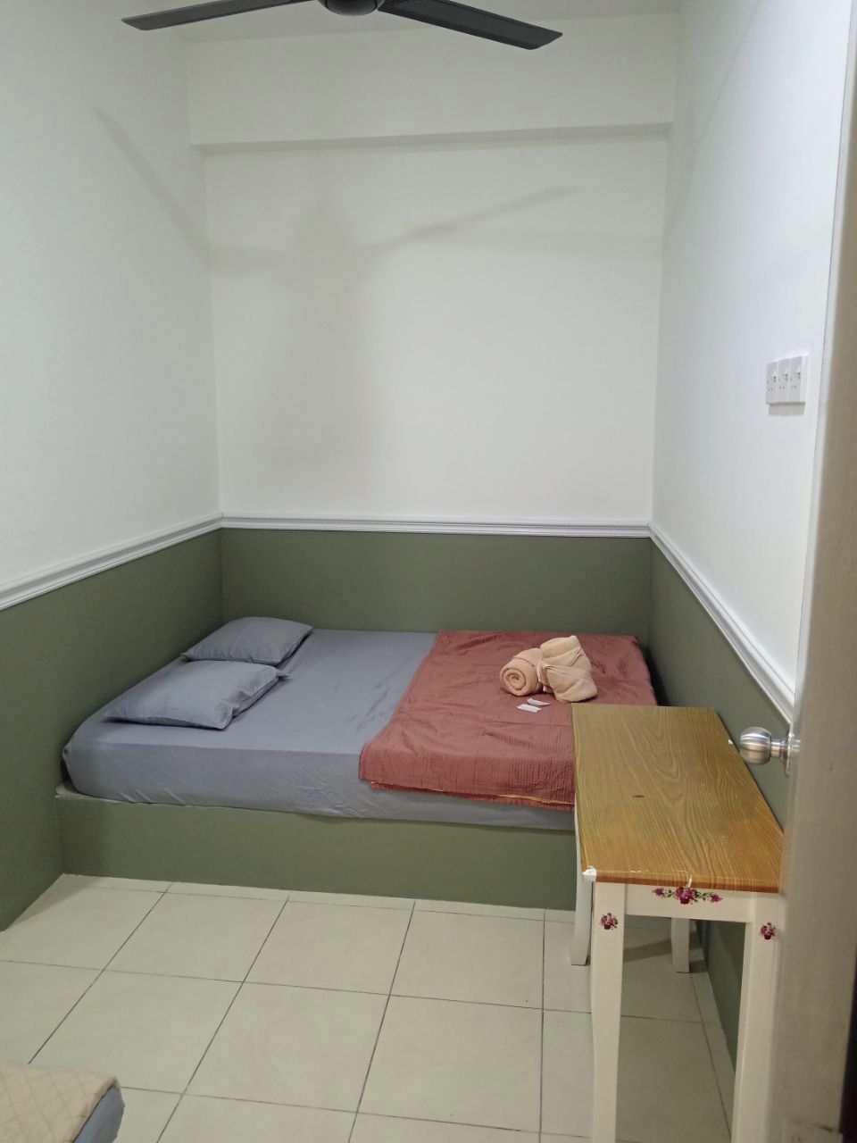 Quarto pequeno com cama de solteiro, decoração em tons de verde e branco, ventilador de teto e mesa de madeira.