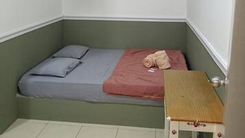 Quarto pequeno com cama de solteiro, decoração em tons de verde e branco, ventilador de teto e mesa de madeira.