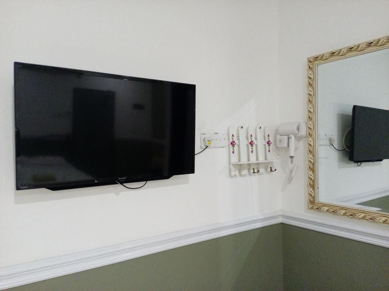 Un televisor montado en la pared, un secador de pelo y un espejo con marco decorativo.