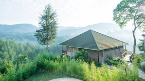 A house with a sloped roof, surrounded by dense greenery and mountains in the background. - Gorilla Leisure Lodge (Bugambira)