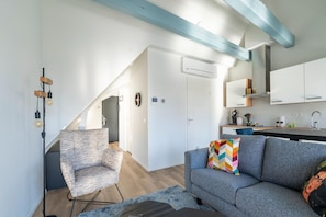 A modern living space with a grey sofa, a patterned armchair, and a colorful pillow. There is a kitchen area with white cabinets and a built-in oven. A floor lamp with a light bulb is visible.